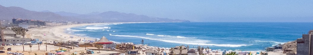 A wide view along Cerritos Beach, lines of surf running toward the point with mountains beyond